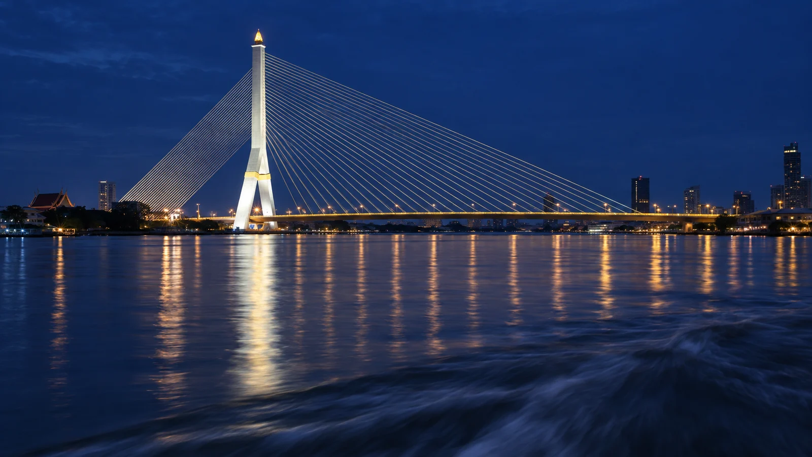 The Rama VIII bridge in Bangkok lit at dusk, seen from the water